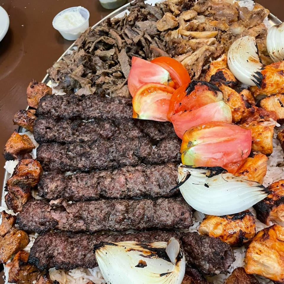 A large platter of Erbil Kabob's grilled meats, including kabobs, chicken, and shawarma, with rice, tomatoes, and onions.